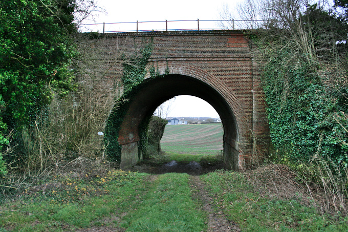 Wide-arched railway bridge under railway line, with grassy track leading to arch, and cultivated fields beyond